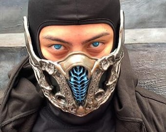 May include: A silver and blue face mask with a detailed design. The mask covers the nose and mouth and has a small opening for the eyes. The mask is worn by a person with blue contact lenses.