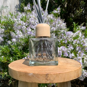 May include: A clear glass reed diffuser bottle with a wooden lid and gray reeds. The bottle has a label that reads "Pure Wight Isle of Wight Reed Diffuser Poured by the Sea".