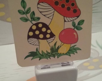 May include: A white night light with a mushroom design. The mushrooms are red and white with black polka dots and yellow stems. The night light has a clear plastic cover and a white base.