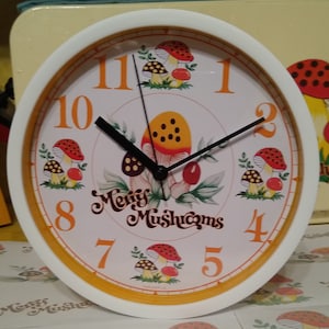 Könnte beinhalten: Eine weiße Uhr mit einem gelben Zifferblatt und schwarzen Zeigern. Die Uhr hat ein Pilz-Motiv mit roten und weißen Pilzen und dem Text "Merry Mushrooms".