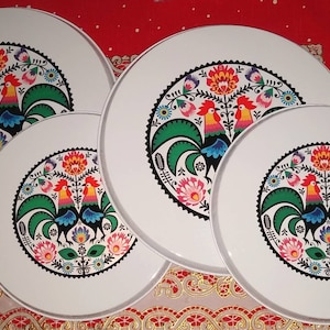 Puede incluir: Bandejas redondas blancas con un diseño colorido de gallos y flores. Los gallos tienen crestas rojas, picos amarillos y plumas verdes. Las bandejas están dispuestas sobre una superficie roja estampada.
