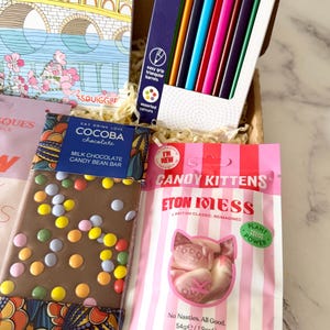 May include: A gift box containing a coloring book with a bridge illustration, a pack of 12 triangular colored pencils, a Cocoba milk chocolate candy bean bar, and a 1.9 ounce bag of Candy Kittens Eton Mess vegan gummy candies.