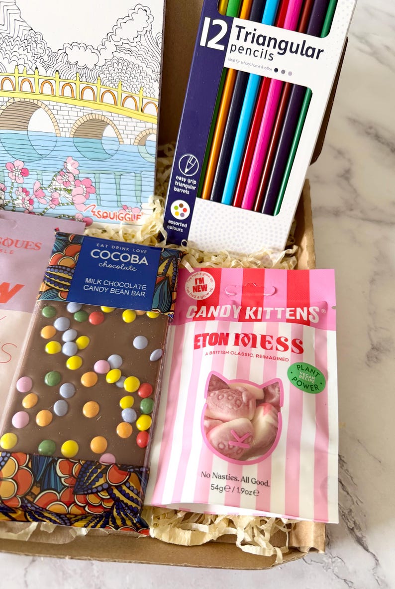 May include: A gift box containing a coloring book with a bridge illustration, a pack of 12 triangular colored pencils, a Cocoba milk chocolate candy bean bar, and a 1.9 ounce bag of Candy Kittens Eton Mess vegan gummy candies.