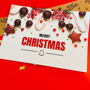 May include: A white rectangular greeting card with the text MERRY CHRISTMAS in black and red letters. The card features dark red glittery ornaments, candy canes, red berries, and small gold stars. A black bell icon is centred below the text.