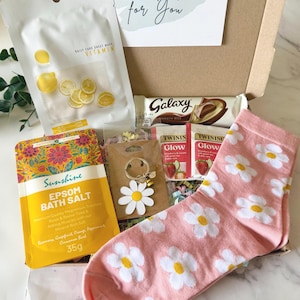 May include: A gift box containing a white vitamin sheet mask, a Galaxy chocolate bar, two Twinings Glow tea bags, a yellow Sunshine Epsom bath salt packet, a daisy keychain with a bee charm, and pink socks with a white daisy pattern.