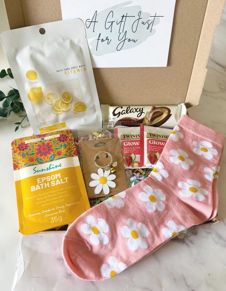 May include: A gift box containing a white vitamin sheet mask, a Galaxy chocolate bar, two Twinings Glow tea bags, a yellow Sunshine Epsom bath salt packet, a daisy keychain with a bee charm, and pink socks with a white daisy pattern.