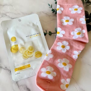 May include: A pink sock with a repeating white and yellow daisy flower pattern alongside a white Eunyul Daily Care Sheet Mask Vitamin packet featuring images of sliced lemons and yellow text on the front of the packaging.