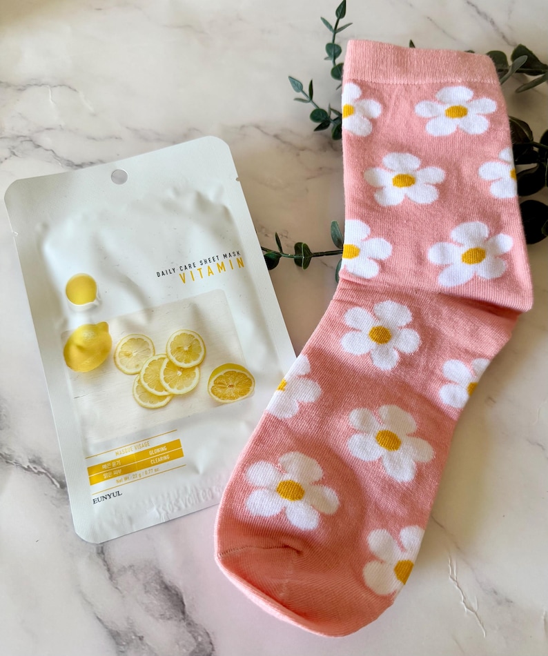 May include: A pink sock with a repeating white and yellow daisy flower pattern alongside a white Eunyul Daily Care Sheet Mask Vitamin packet featuring images of sliced lemons and yellow text on the front of the packaging.