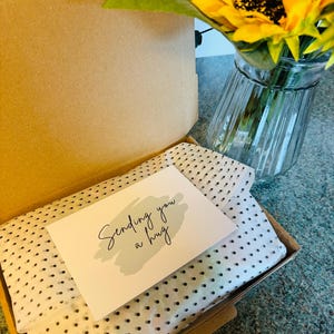 May include: An open cardboard box with a white and black polka dot tissue paper lining. A card with the words "Sending you a hug" is on top. A sunflower in a glass vase is in the background.