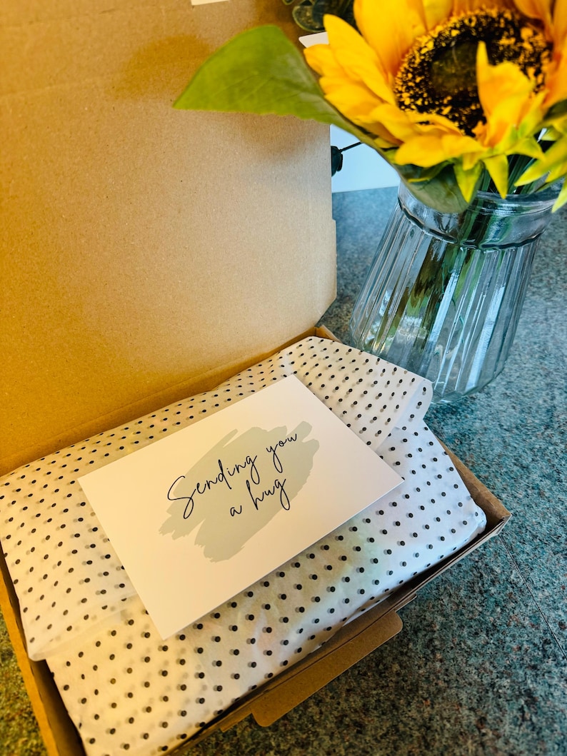 May include: An open cardboard box with a white and black polka dot tissue paper lining. A card with the words "Sending you a hug" is on top. A sunflower in a glass vase is in the background.