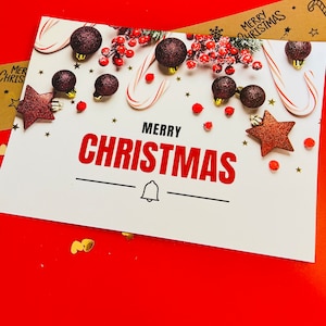 May include: A white rectangular greeting card with the text MERRY CHRISTMAS in black and red letters. The card features dark red glittery ornaments, candy canes, red berries, and small gold stars. A black bell icon is centered below the text.