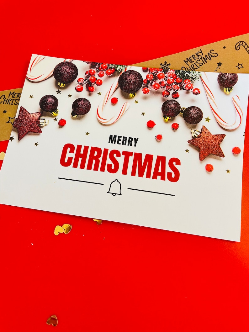 May include: A white rectangular greeting card with the text MERRY CHRISTMAS in black and red letters. The card features dark red glittery ornaments, candy canes, red berries, and small gold stars. A black bell icon is centered below the text.