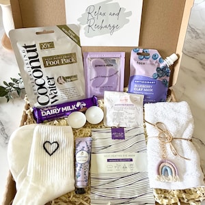 May include: A gift box containing a coconut foot pack, purple eye masks, a blueberry clay mask, a Cadbury Dairy Milk chocolate bar, two white tea light candles, a lavender bath bomb, white socks with a black heart, lavender hand cream, a self-heating eye mask, and a white towel with a rainbow keychain.
