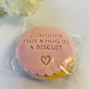 May include: A round biscuit with a scalloped edge, topped with a smooth pink fondant layer. The text "CONSIDER THIS A HUG IN A BISCUIT" is embossed in the centre above a small heart shape. The biscuit is sealed inside a clear plastic wrapper.