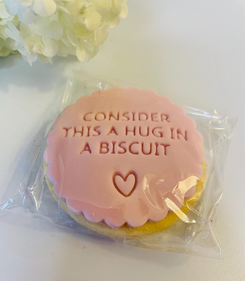 May include: A round biscuit with a scalloped edge, topped with a smooth pink fondant layer. The text "CONSIDER THIS A HUG IN A BISCUIT" is embossed in the centre above a small heart shape. The biscuit is sealed inside a clear plastic wrapper.