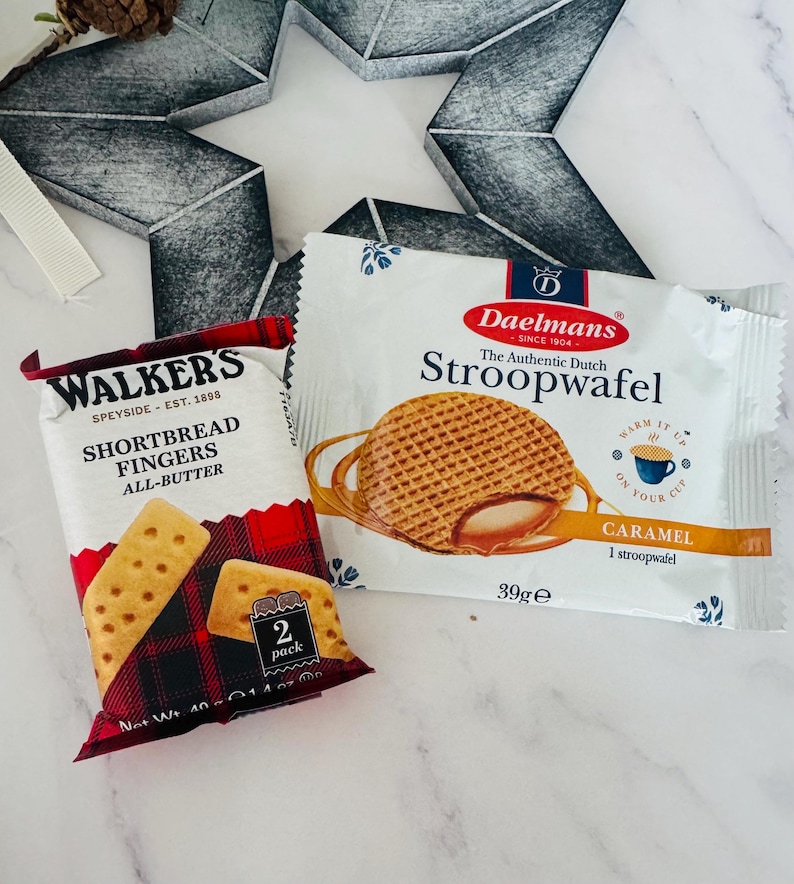 May include: A package of Walkers Shortbread Fingers with a red and black plaid design and a package of Daelmans Stroopwafel featuring a round caramel-filled waffle graphic and the text Warm It Up On Your Cup, both 39g and 40g net weight.