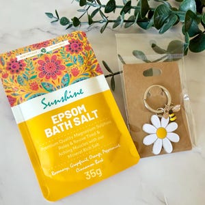 May include: A yellow pouch of Sunshine Epsom Bath Salt with a floral pattern, containing 35g of magnesium sulphate, rosemary, grapefruit, orange, peppermint, and cinnamon bark. Next to it is a daisy and bee metal keychain on a brown card.