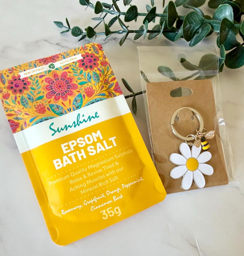 May include: A yellow pouch of Sunshine Epsom Bath Salt with a floral pattern, containing 35g of magnesium sulphate, rosemary, grapefruit, orange, peppermint, and cinnamon bark. Next to it is a daisy and bee metal keychain on a brown card.