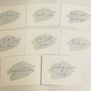 May include: A collection of eight rectangular white greeting cards, each featuring a light grey brushstroke background and black cursive text. The phrases include Thinking of You, Good Luck, Relax and Recharge, A Gift Just for You, Get Well Soon, Thank you, Congratulations, and Sending you a hug.