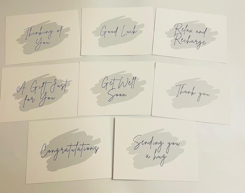 May include: A collection of eight rectangular white greeting cards, each featuring a light grey brushstroke background and black cursive text. The phrases include Thinking of You, Good Luck, Relax and Recharge, A Gift Just for You, Get Well Soon, Thank you, Congratulations, and Sending you a hug.