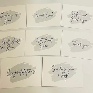 May include: A collection of white greeting cards with various messages in elegant script. Each card features a gray brushstroke design. Messages include "Thinking of You," "Good Luck," and "Relax and Recharge."