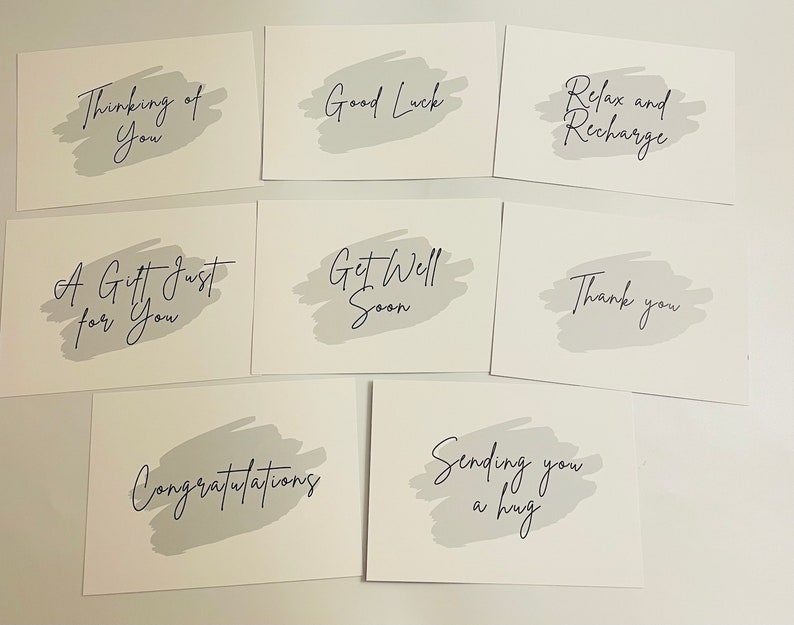 May include: A collection of white greeting cards with various messages in elegant script. Each card features a gray brushstroke design. Messages include "Thinking of You," "Good Luck," and "Relax and Recharge."