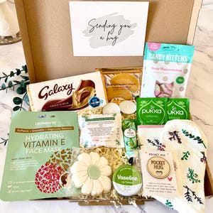 May include: A cardboard gift box containing a Galaxy chocolate bar, Candy Kittens sour watermelon sweets, a DermaV10 Vitamin E face mask, a flower-shaped cookie, Vaseline lip therapy, Pukka tea bags, socks, and a wooden pocket hug token.