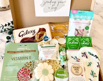 May include: A cardboard gift box containing a Galaxy chocolate bar, Candy Kittens sour watermelon sweets, a DermaV10 Vitamin E face mask, a flower-shaped cookie, Vaseline lip therapy, Pukka tea bags, socks, and a wooden pocket hug token.