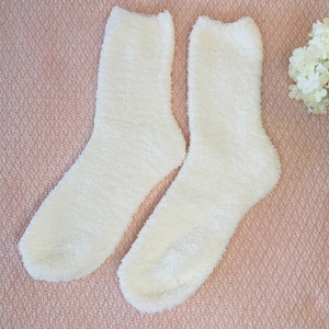 May include: A pair of fluffy, off-white socks. The socks are made of a soft, textured material and are designed to be cozy. The socks are displayed on a pink surface, with a few white flowers in the background.