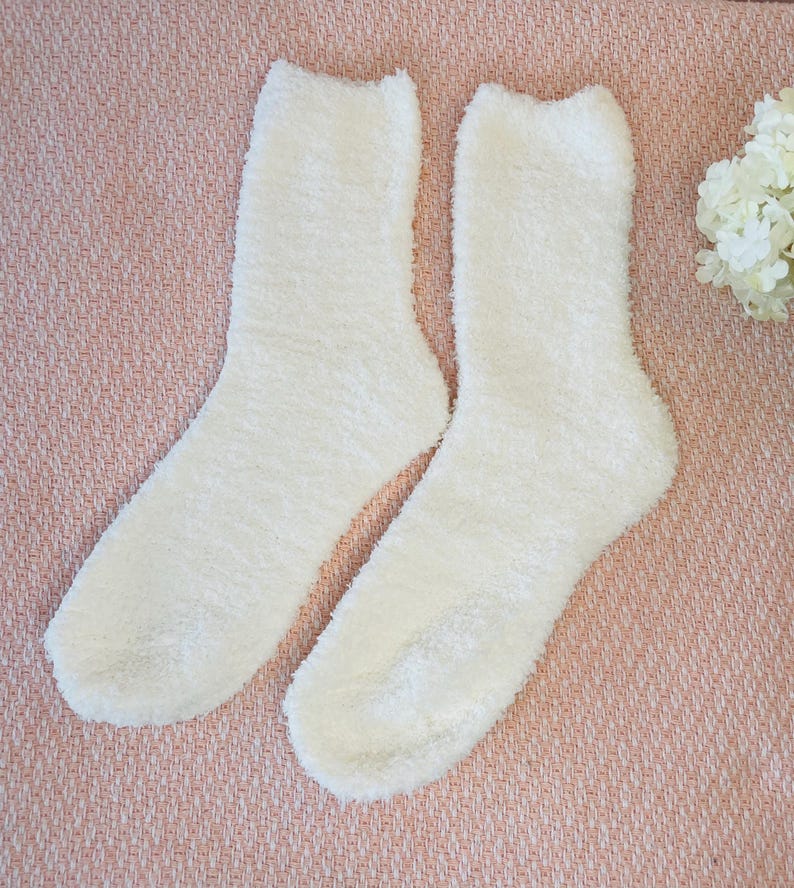 May include: A pair of fluffy, off-white socks. The socks are made of a soft, textured material and are designed to be cozy. The socks are displayed on a pink surface, with a few white flowers in the background.