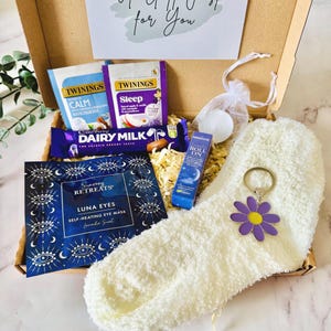 May include: A gift box filled with self-care items. Includes Twinings tea bags, a Cadbury Dairy Milk chocolate bar, a Luna Eyes self-heating eye mask, a roll-on, a white fuzzy sock, and a daisy keychain. The box has a card that says "A Gift Just for You".