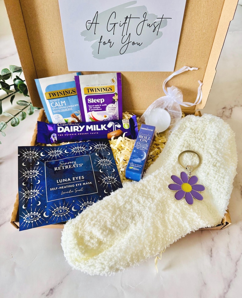 May include: A gift box filled with self-care items. Includes Twinings tea bags, a Cadbury Dairy Milk chocolate bar, a Luna Eyes self-heating eye mask, a roll-on, a white fuzzy sock, and a daisy keychain. The box has a card that says "A Gift Just for You".