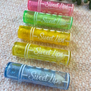 May include: Five cylindrical lip balm tubes arranged vertically, each featuring a translucent coloured plastic casing in pink, green, yellow, light yellow, and blue. Each tube is printed with the white text Sweet Lips BY BEAUTY 4 BRITAIN.