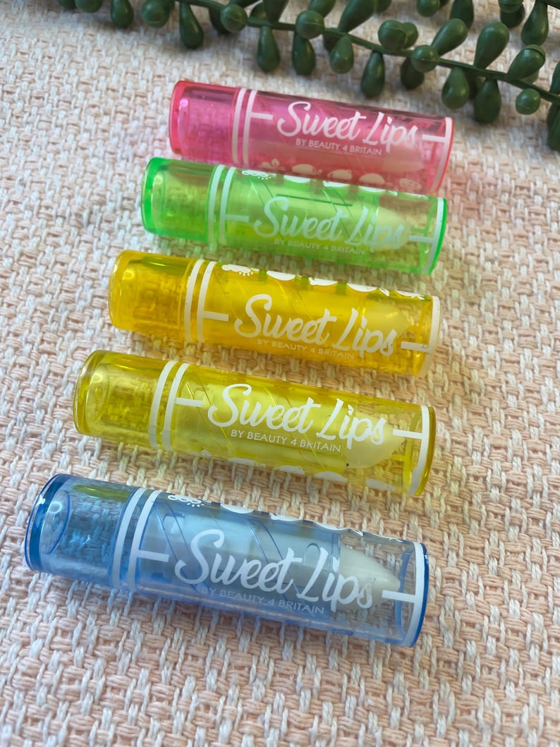 May include: Five cylindrical lip balm tubes arranged vertically, each featuring a translucent coloured plastic casing in pink, green, yellow, light yellow, and blue. Each tube is printed with the white text Sweet Lips BY BEAUTY 4 BRITAIN.