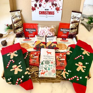 May include: A cardboard gift box containing two pairs of green socks with gingerbread men and candy cane patterns, two Galaxy hot chocolate sachets, two Yorkshire Tea bags, a bag of marshmallows, two Daelmans stroopwafels, two Walkers shortbread packs, two Santa chocolates, and a Merry Christmas card.