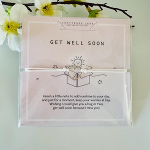 May include: A 'Get Well Soon' card with a gold-colored heart-shaped charm and a small drawing of a sun. The card has text that reads, "Here's a little note to add sunshine to your day..." and is packaged in clear plastic.
