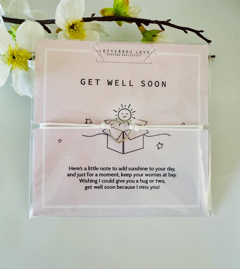 May include: A 'Get Well Soon' card with a gold-colored heart-shaped charm and a small drawing of a sun. The card has text that reads, "Here's a little note to add sunshine to your day..." and is packaged in clear plastic.