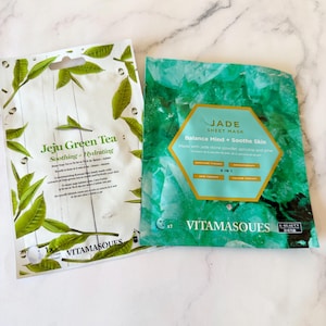 May include: Two rectangular face mask packets. The left packet is white with green tea leaf illustrations and text reading Jeju Green Tea Soothing + Hydrating. The right packet is green with a jade stone pattern and text reading Jade Sheet Mask.