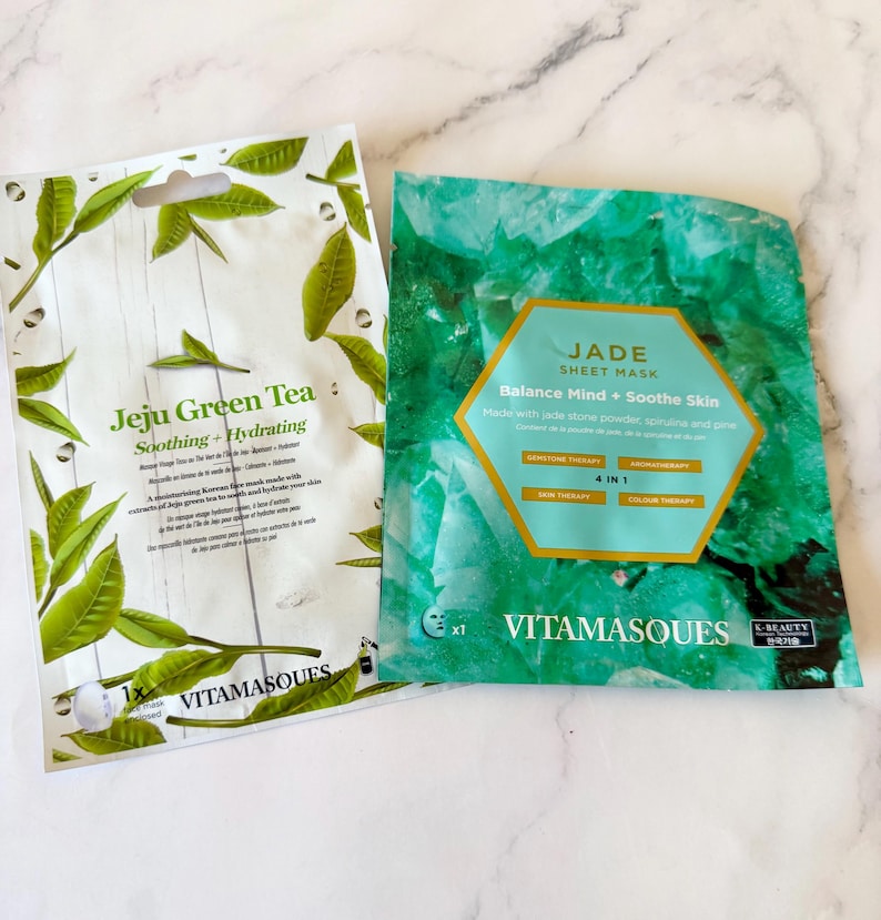 May include: Two rectangular face mask packets. The left packet is white with green tea leaf illustrations and text reading Jeju Green Tea Soothing + Hydrating. The right packet is green with a jade stone pattern and text reading Jade Sheet Mask.