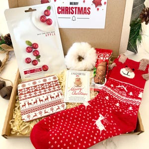 May include: A Christmas gift box containing a white apple sheet mask, a red and white patterned coaster, a white faux fur scrunchie, a KitKat chocolate bar, a small card with a gingerbread man, and a red sock with a reindeer pattern.