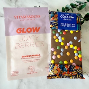 May include: A Vitamasques Glow Juicy Berries biodegradable sheet mask in a light purple and cream pouch next to a Cocoba milk chocolate candy bean bar with colorful candy pieces in a blue and patterned wrapper with the text Eat Drink Love.