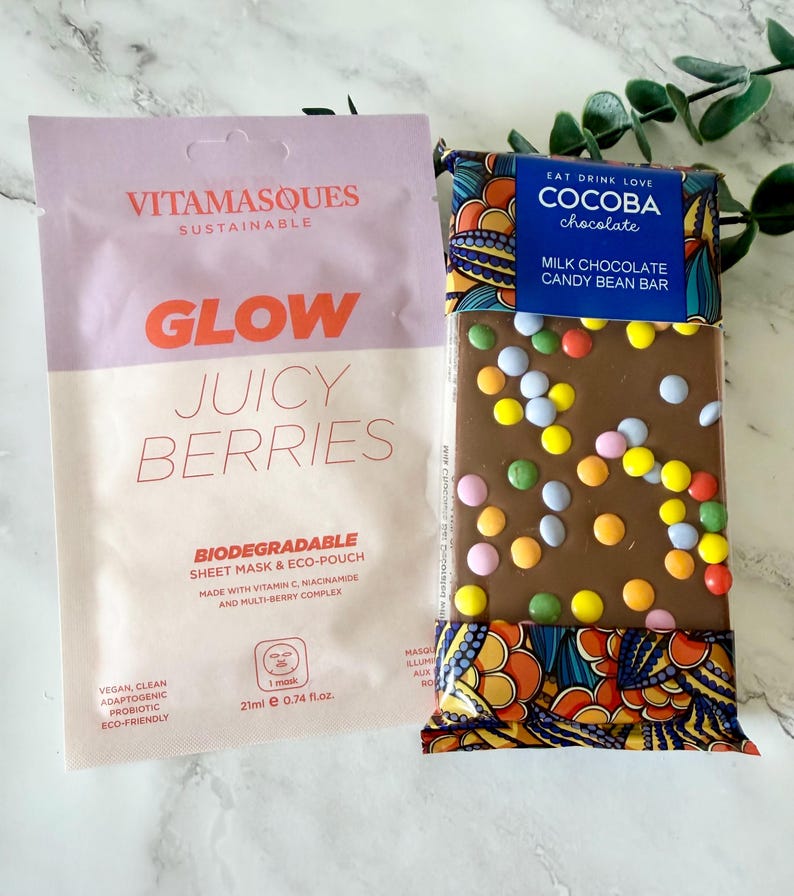 May include: A Vitamasques Glow Juicy Berries biodegradable sheet mask in a light purple and cream pouch next to a Cocoba milk chocolate candy bean bar with colorful candy pieces in a blue and patterned wrapper with the text Eat Drink Love.