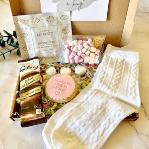 May include: A gift box containing a white cable knit sock, a packet of Galaxy instant hot chocolate, a Sensory Retreats self-heating eye mask, a bag of pink and white marshmallows, and a round biscuit with pink icing reading Consider this a hug in a biscuit.