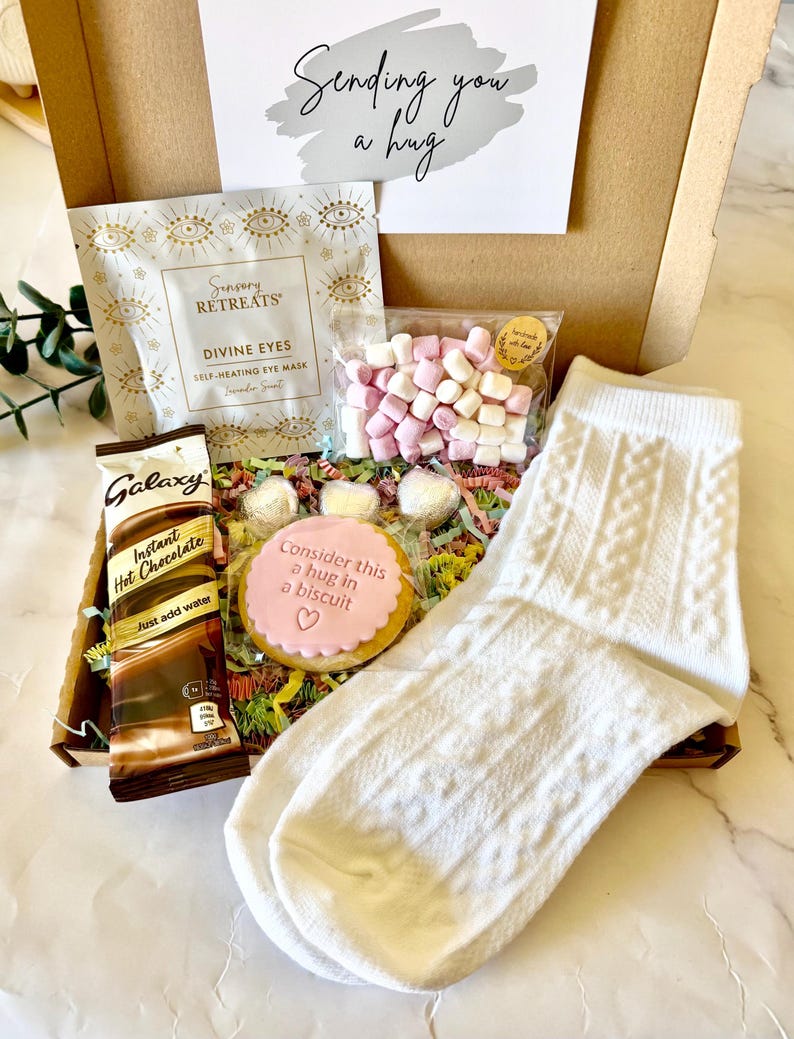 May include: A gift box containing a white cable knit sock, a packet of Galaxy instant hot chocolate, a Sensory Retreats self-heating eye mask, a bag of pink and white marshmallows, and a round biscuit with pink icing reading Consider this a hug in a biscuit.