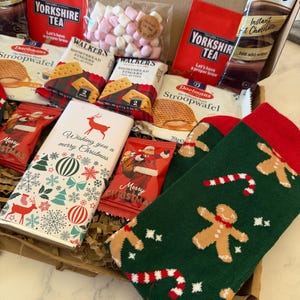 May include: A gift box containing two packs of Yorkshire Tea, two Daelmans Stroopwafels, two Walkers Shortbread Fingers, a Galaxy hot chocolate sachet, a bag of pink and white marshmallows, a chocolate bar with a reindeer, and green socks with gingerbread men.