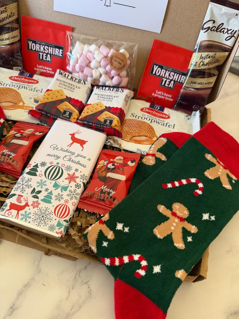 May include: A gift box containing two packs of Yorkshire Tea, two Daelmans Stroopwafels, two Walkers Shortbread Fingers, a Galaxy hot chocolate sachet, a bag of pink and white marshmallows, a chocolate bar with a reindeer, and green socks with gingerbread men.