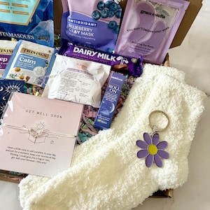 May include: A gift box filled with self-care items. Includes a face mask, eye masks, tea bags, a chocolate bar, a roll-on, a pair of fuzzy socks, and a daisy keychain. The box also contains a card that says "Get Well Soon."