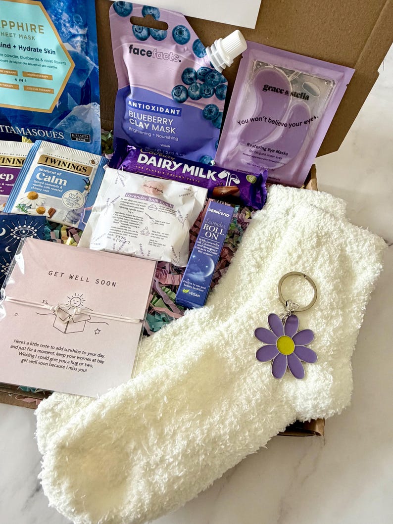 May include: A gift box filled with self-care items. Includes a face mask, eye masks, tea bags, a chocolate bar, a roll-on, a pair of fuzzy socks, and a daisy keychain. The box also contains a card that says "Get Well Soon."