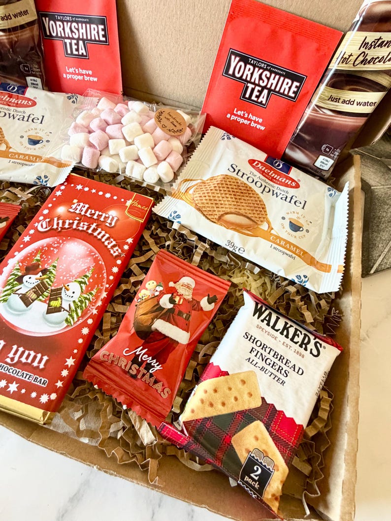 May include: A gift box filled with assorted treats including two red Taylors of Harrogate Yorkshire Tea packets, a Daelmans caramel stroopwafel, a Walkers shortbread fingers pack, a Merry Christmas chocolate bar, a Santa snack bag, and marshmallows.