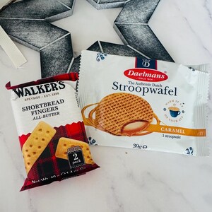May include: A packet of Walkers Shortbread Fingers with a red and black tartan pattern and a packet of Daelmans Stroopwafel featuring a round caramel-filled waffle graphic and the text Warm It Up On Your Cup, both 39g and 40g net weight.